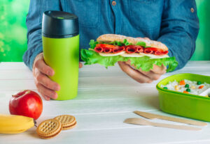 Männliche Hände halten Sandwich und Thermoskanne während des Essens sein Mittagessen aus Lunchbox auf weißem hölzernen Hintergrund